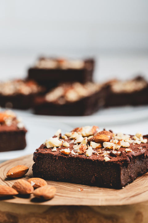A sugar free keto brownie with a crushed almond topping on a wooden tray with almonds scattered around and more brownies in the background