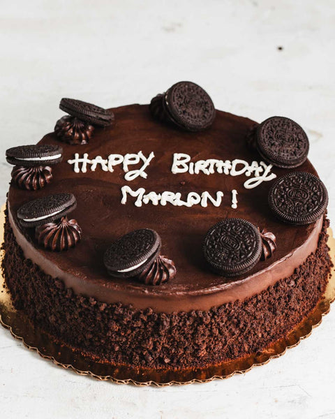 A chocolate frosted cake with chocolate frosting dollops and oreos as decoration, rolled in chocolate cake crumbs. "Happy Birthday Marlon!" is written on top.