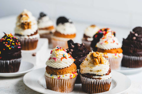 White plates of chocolate, vanilla, and banana walnut cupcakes with chocolate and vanilla frosting topped with rainbow sprinkles, walnuts, chocolate chips, and oreo cookies.
