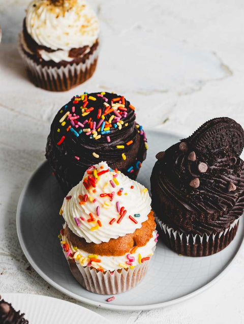 A close up of a plate of three cupcakes: A vanilla cupcake with vanilla icing and rainbow sprinkles, a chocolate cupcake with chocolate icing, chocolate chips, and an oreo cookie, and a chocolate cupcake with chocolate icing and rainbow sprinkles. Other cupcakes are around them.