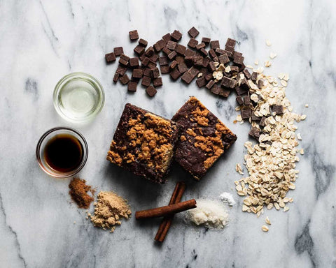 Two oatmeal brookies on a marble surface surrounded by chocolate chunks, oats, vanilla extract, oil, cinnamon sticks, ground cinnamon, brown sugar, and flour.