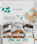 An open white gift box with colorful floral patters displaying an assortment of packaged food items, including an oatmeal brookie, an apricot almont bar, a walnut chocolate brownie, a german chocolate brownie, an oatmeal chocolate cookie, and a cookie crumble cookie