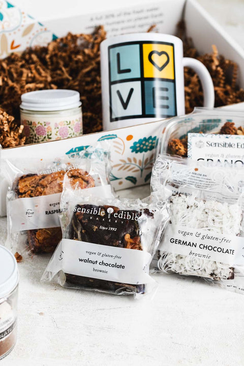 Gift box with contents partially taken out in front of it: Jar of hot chocolate mix, walnut chocolate brownie, sensible edibles mug, candle, granola, and german chocolate brownie.