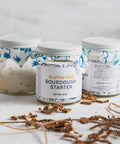 Three jars of gluten-free sourdough starter on a white table with decorative fabric lids.