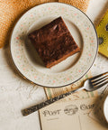 Chocolate chunk brownie on a decorative plate with a fork and postcard in the background