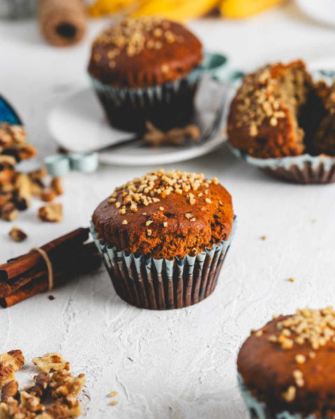 Close-up of a vegan and gluten-free banana walnut muffin topped with chopped walnuts, surrounded by more muffins on a light textured surface.