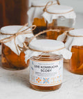 Jars of kombucha with a live scoby, labeled and with cloth covers tied with twine, on a kitchen counter.