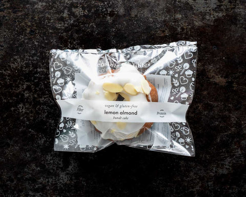 Packaged mini lemon almond bundt cake with white icing and almond slices on a dark surface.