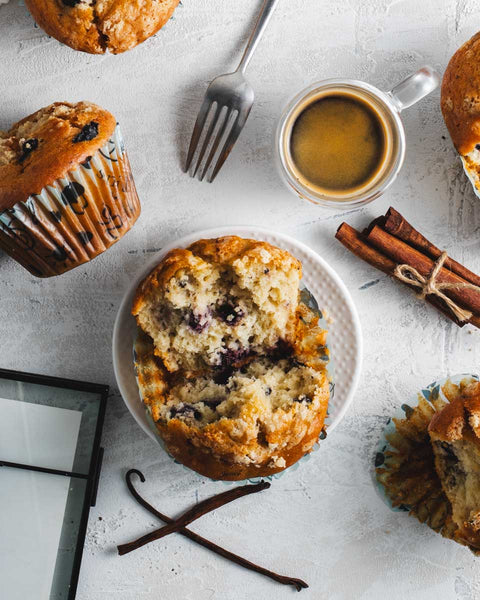 Top view of a blueberry crumb muffin broken in half, revealing the moist interior, set on a small plate surrounded by a cup of coffee, cinnamon sticks, vanilla beans on a light textured background.