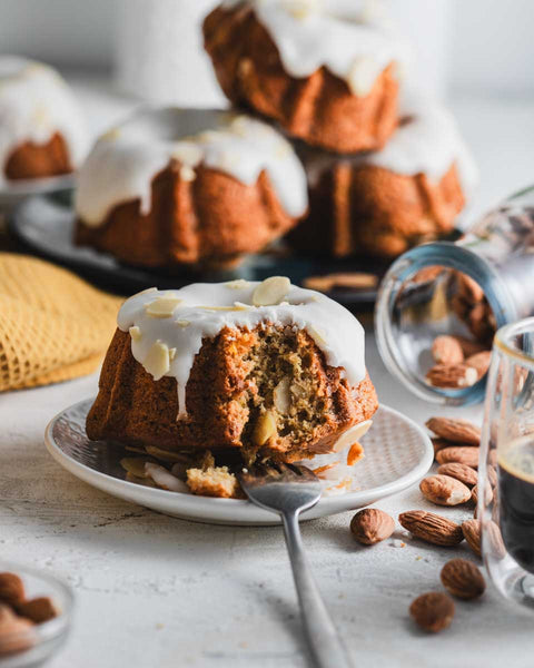 Mini lemon almond bundt cake with white icing and almond slices on a plate, surrounded by whole almonds and a glass of espresso, with more cakes in the background.
