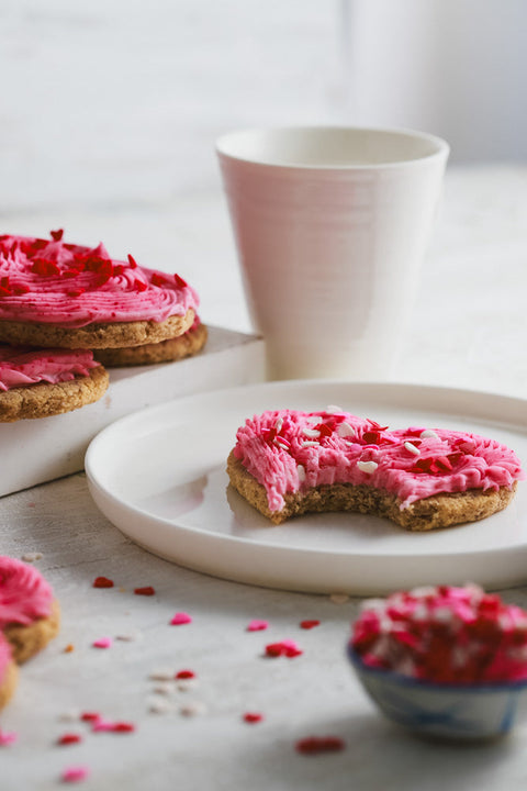 Heart-shaped cookie with pink frosting and red sprinkles, a bite taken out, on a white plate with a white cup in the background.