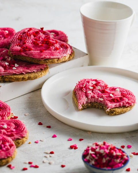 Heart-shaped cookie with pink frosting and red sprinkles, a bite taken out, on a white plate, with more cookies, a white cup, and a bowl of sprinkles.