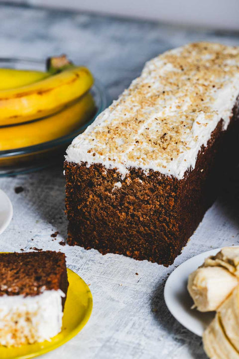 A banana walnut loaf cake with a slice cut out, alongside fresh bananas and walnuts on a grey background.