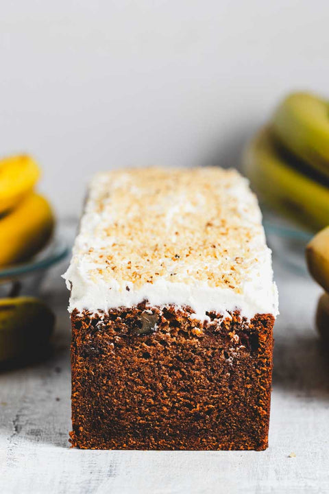A banana walnut loaf cake with a slice cut out, alongside fresh bananas and walnuts on a grey background.