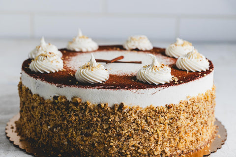 A side view of a cake with white frosting dusted with cooca powder and rolled in crushed nuts with white icing and cinnamon stick decoration