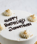 A close up of the top of a vanilla frosted cake with "happy birthday jonathan" written in brown icing and decorative dollops of white icing topped with a crushed nut garnish. The side of the cake is rolled in crushed nuts.