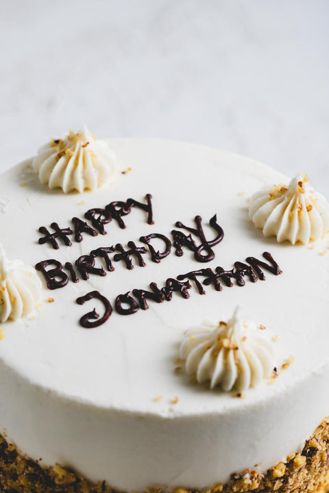 A close up of the top of a vanilla frosted cake with "happy birthday jonathan" written in brown icing and decorative dollops of white icing topped with a crushed nut garnish. The side of the cake is rolled in crushed nuts.