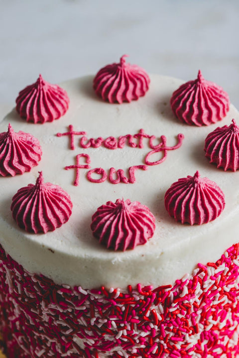 A decorated cake with the words 'Twenty four' written on it in icing, surrounded by pink frosting dollops on a white background. The side of the cake is rolled in pink and white sprinkles.