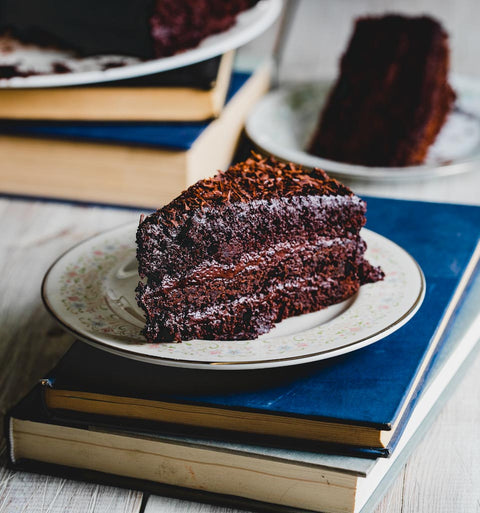 Slice of chocolate fudge cake on a decorative plate, set on a stack of books with more slices in the background.