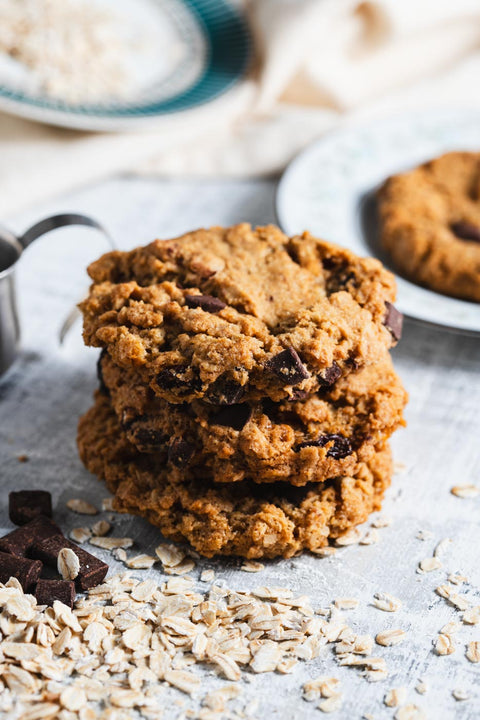 Stack of oatmeal chocolate chunk cookies with visible oats and chocolate pieces, on a light surface with a plate of cookies in the background.