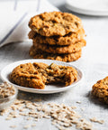 Stack of apple oatmeal cookies with one cookie on a plate with a bite taken out of it, showcasing the texture and apples inside. The cookies are on a light background with scattered oats around.