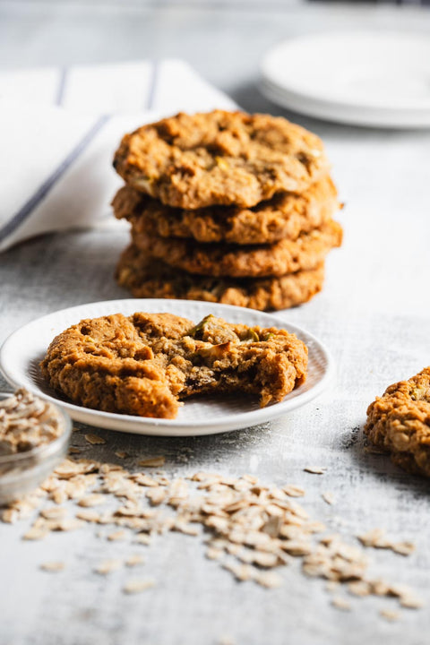 Stack of apple oatmeal cookies with one cookie on a plate with a bite taken out of it, showcasing the texture and apples inside. The cookies are on a light background with scattered oats around.