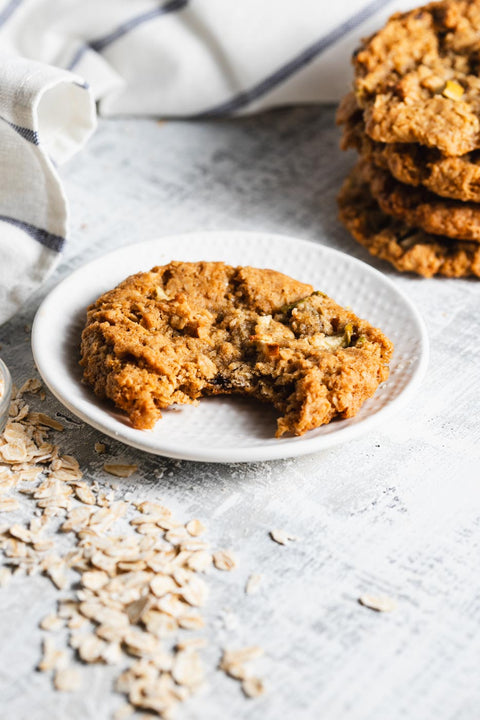 An apple oatmeal cookie on a white plate with a bite taken out of it. There is a stack of more cookies behing it and oats piled around.