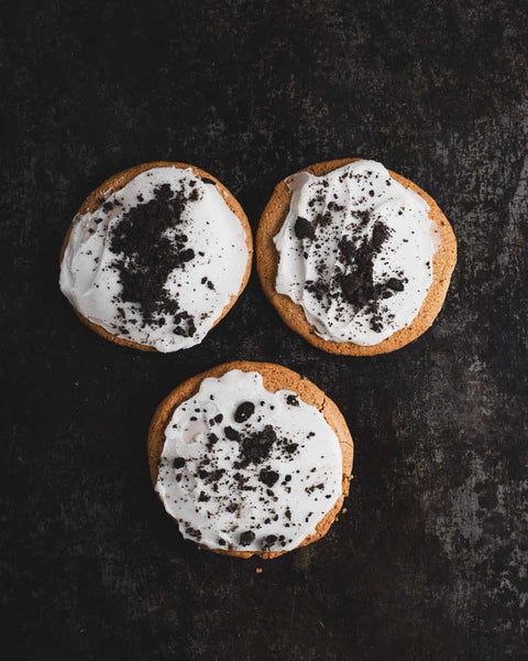 Three frosted cookie crunches with white icing and crushed vegan Oreos, arranged on a dark background.