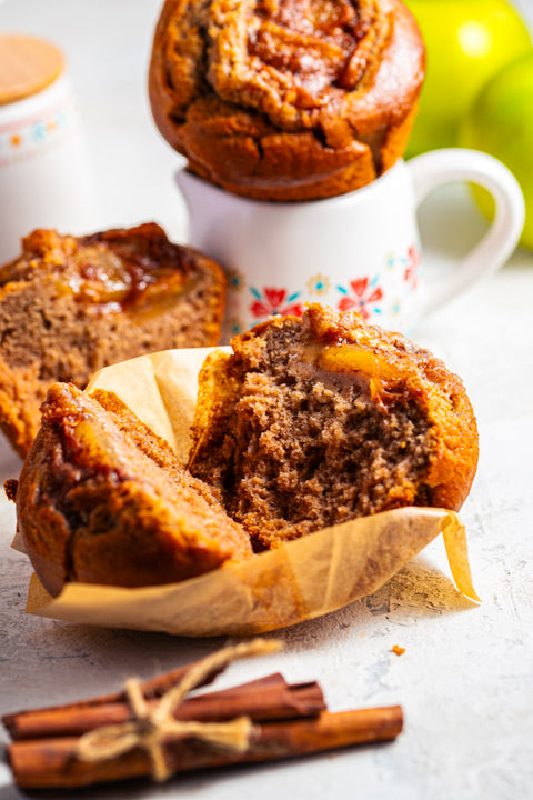 Apple pie muffin cut in half with a whole muffin, cinnamon sticks, and an apple in the background.