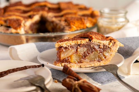Slice of apple pie on a plate with the rest of the pie in the background and plates and cinnamon sticks around it