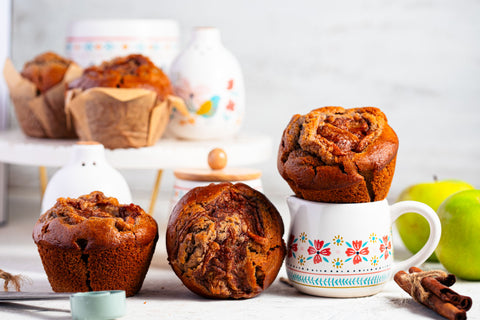 Apple pie muffins on a white surface with a floral mug and apples and cinnamon sticks in the background