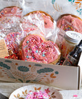Valentine's gift box of sweetheart cookies,pretty in pink cookies, a candle, soap, and body oil with tea cupson a table.