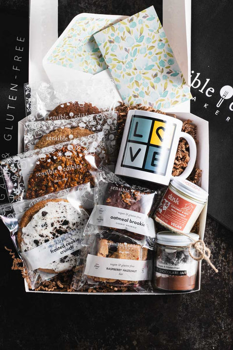 Warmth & Wellness gift box with assorted cookies, a 'LOVE' mug, oatmeal brookie, raspberry hazelnut bar, chocolate jar, vegan candle, and a floral card.