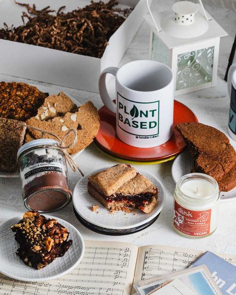 Warmth & Wellness gift box contents displayed, including Sensible Edibles treats, 'Plant Based' mug, chocolate jar, and a vegan candle on a table.