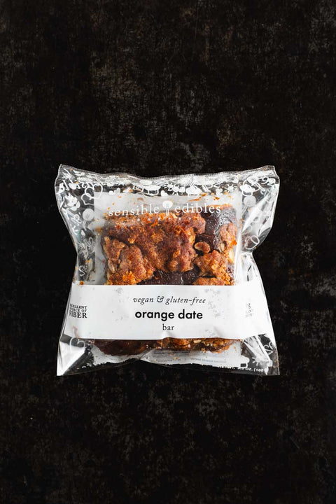 Packaged orange date bar from Sensible Edibles Bakery, labeled as vegan and gluten-free, on a dark background.