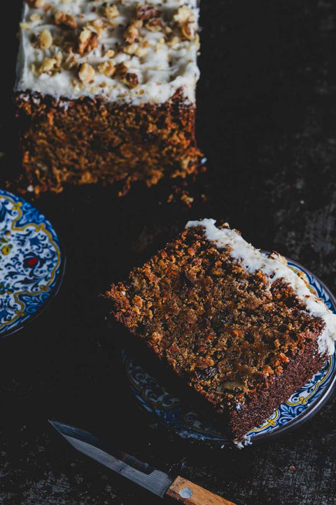 Carrot walnut loaf topped with creamy frosting and crushed walnuts, with a slice cut and served on a decorative plate, showcasing its moist texture.