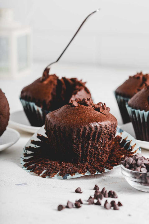 Close-up of a cocoa bean muffin with chocolate chips, unwrapped and placed on a white surface with more muffins in the background.