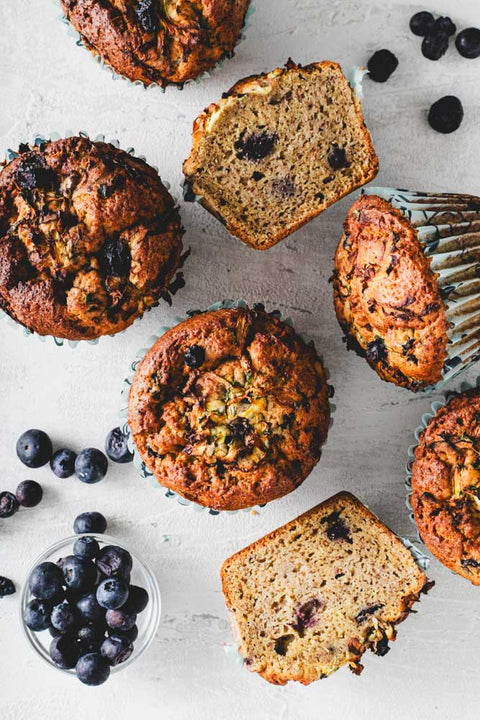 Top view of lemon zucchini protein muffins on a white surface. Some muffins are whole, while others are cut in half to reveal a moist, crumbly texture filled with blueberries.