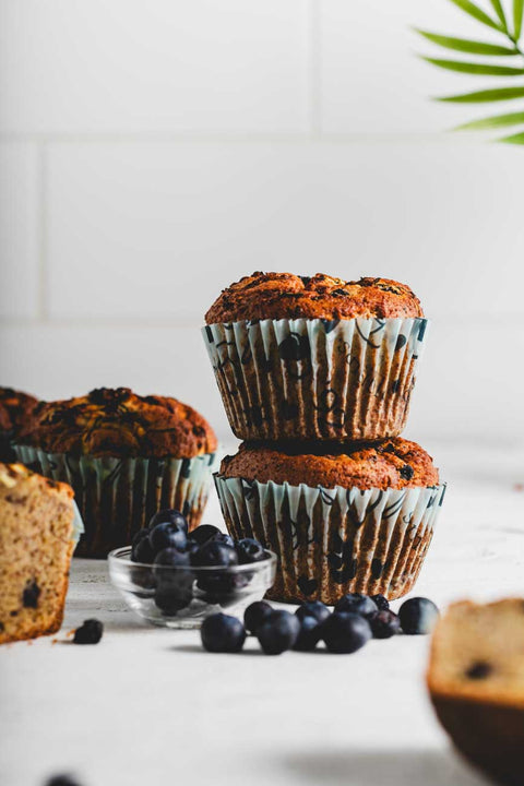 Two stacked lemon zucchini protein muffins in paper liners, with fresh blueberries in a glass bowl and scattered around the muffins.