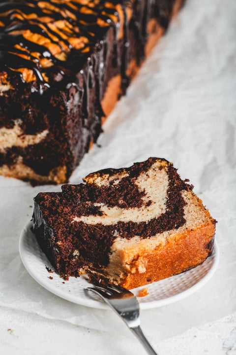 A slice of marble loaf cake on a white plate, showcasing the intricate swirl pattern of chocolate and vanilla, with the rest of the loaf visible in the background.
