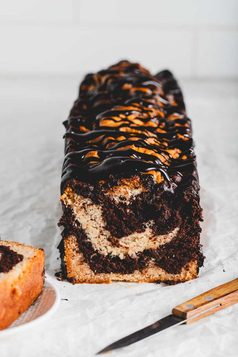 A front view of a whole marble loaf cake, highlighting the marbled chocolate and vanilla interior, topped with a generous drizzle of dark chocolate ganache.