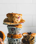 Stack of cinnamon pumpkin muffins with cranberries, showing one partially unwrapped muffin on top and a bitten muffin on the side, placed on a white surface.