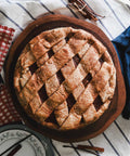Whole apple pie with a lattice crust on a wooden board, surrounded by a striped cloth, plates, and cinnamon sticks.