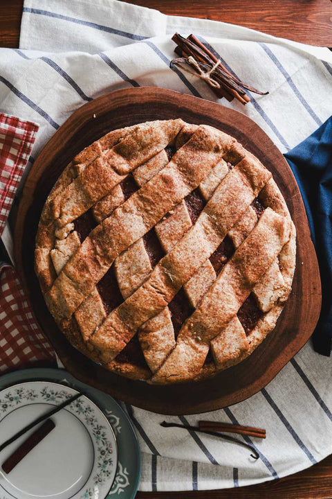Whole apple pie with a lattice crust on a wooden board, surrounded by a striped cloth, plates, and cinnamon sticks.