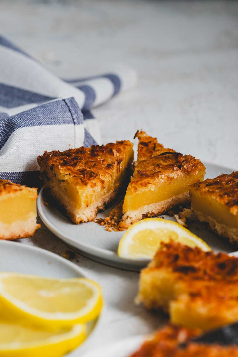 Lemon bars with golden-brown toasted coconut shavings on top scattered around a place with slices of lemon