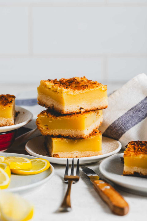 Stack of three lemon bars with golden-brown toasted coconut shavings on top, placed on a white plate with a fork beside it, lemon slices in the background.