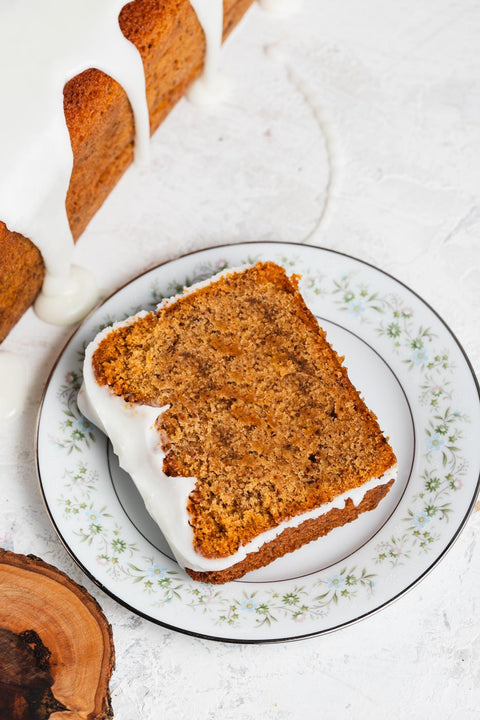 A slice of lemon pound cake with glaze on a plate, with the rest of the cake behind it
