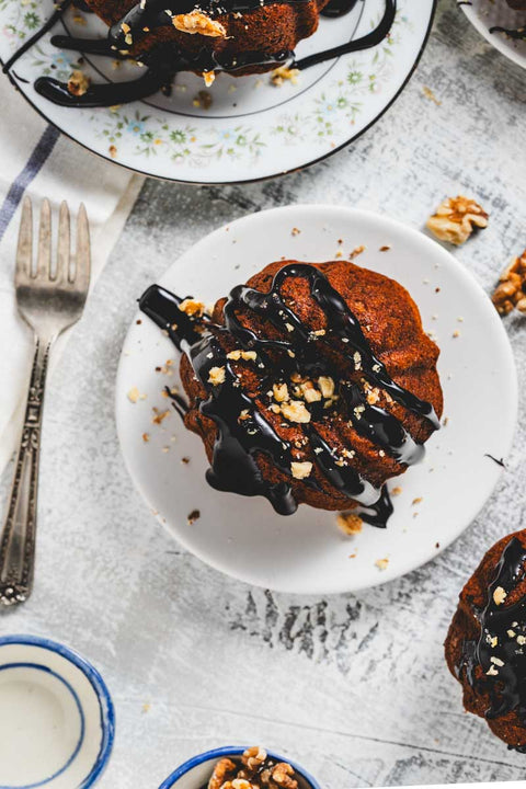 A mini banana bundt cake with chocolate glaze and nuts on top, served on a white plate with a fork and spoon beside it.
