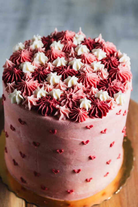 A layered pink velvet cake with white and red frosting decorations, placed on a wooden stand.
