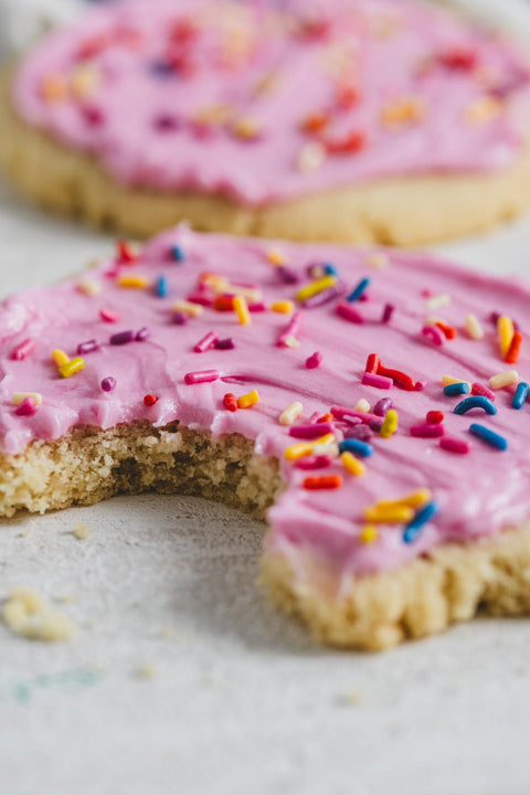 A close up of a sugar cookie with pink icing and rainbow sprinkles with a bite taken out of it on a white background